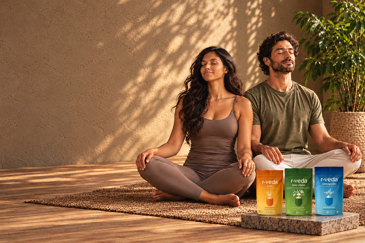 Two people meditating cross-legged in warm sunlight beside r-veda Amla, Baba’s Blend and Ashwagandha pouches on a stone plinth